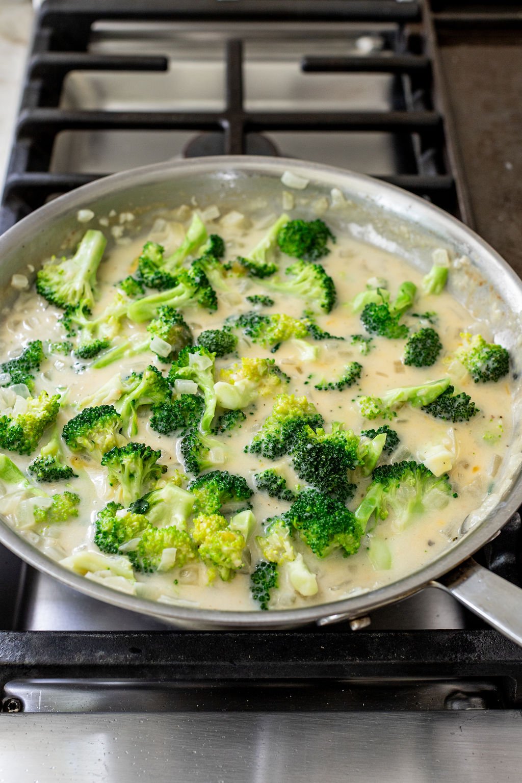 Broccoli florets cooked in a creamy white sauce with chopped onions in a large skillet on a stovetop.