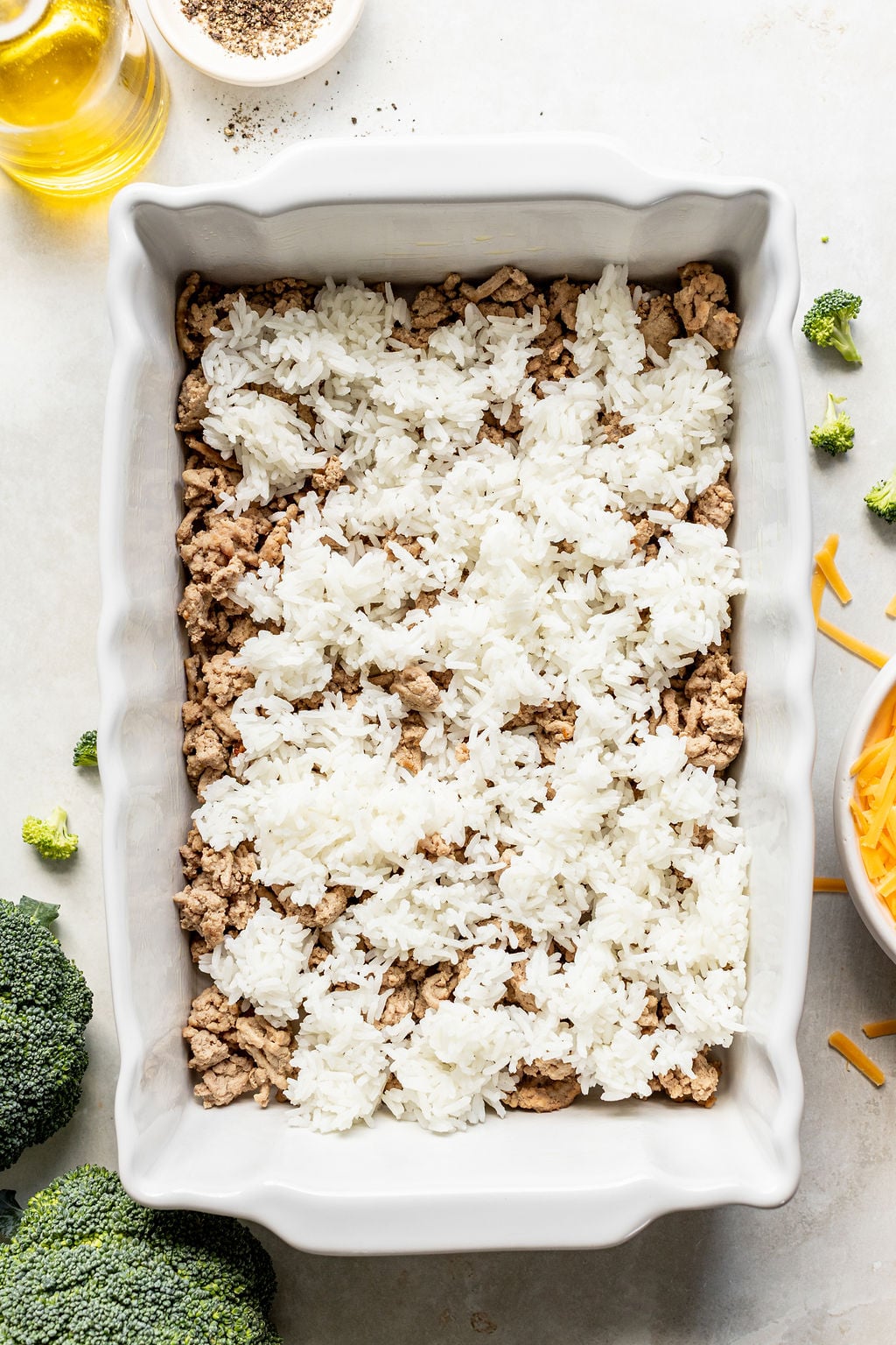 A white baking dish containing a layer of cooked ground meat topped with rice. Broccoli florets, shredded cheese, oil, and pepper are nearby on the countertop.