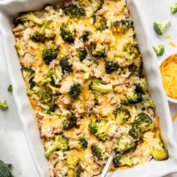 A baked casserole in a white dish, filled with broccoli, rice, cheese, and ground meat, with a serving spoon resting inside. Shredded cheese and broccoli florets are scattered nearby.