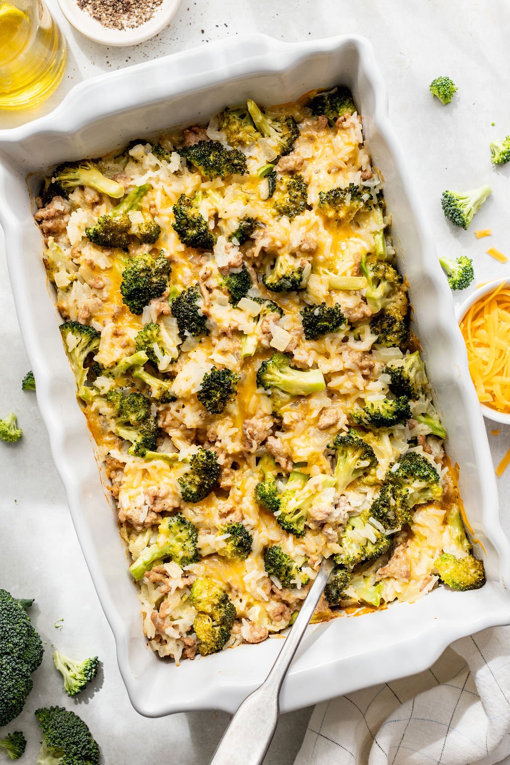 A baked casserole in a white dish, filled with broccoli, rice, cheese, and ground meat, with a serving spoon resting inside. Shredded cheese and broccoli florets are scattered nearby.