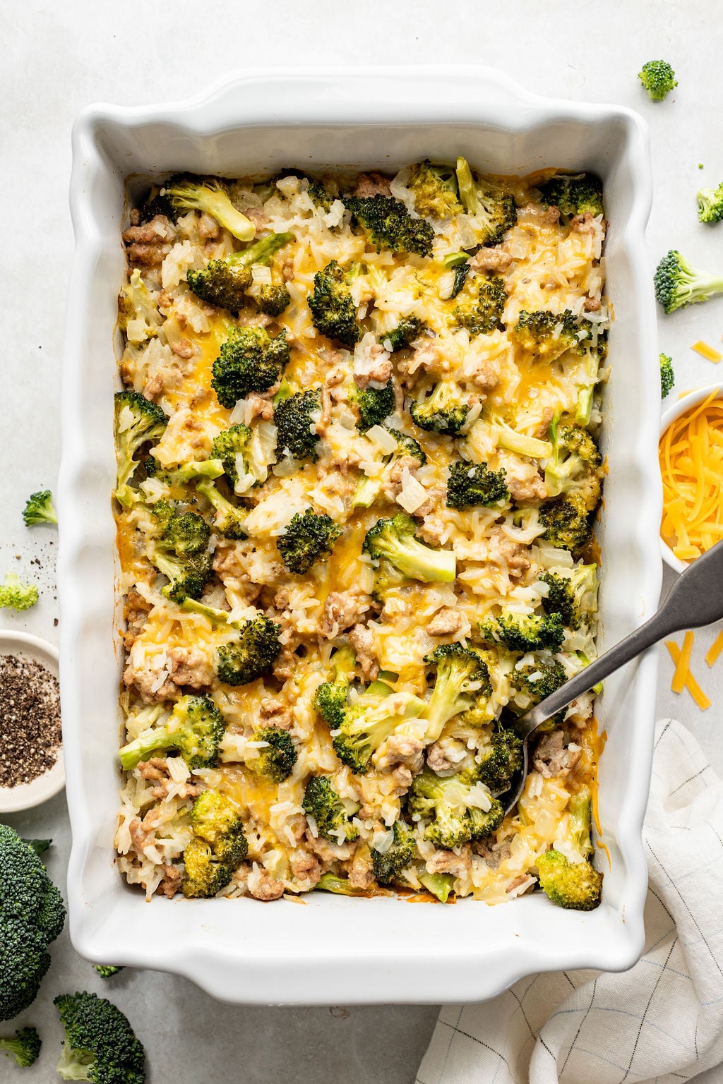 A baked casserole in a white dish containing broccoli, rice, cheese, and ground meat, with a serving spoon on the right side.