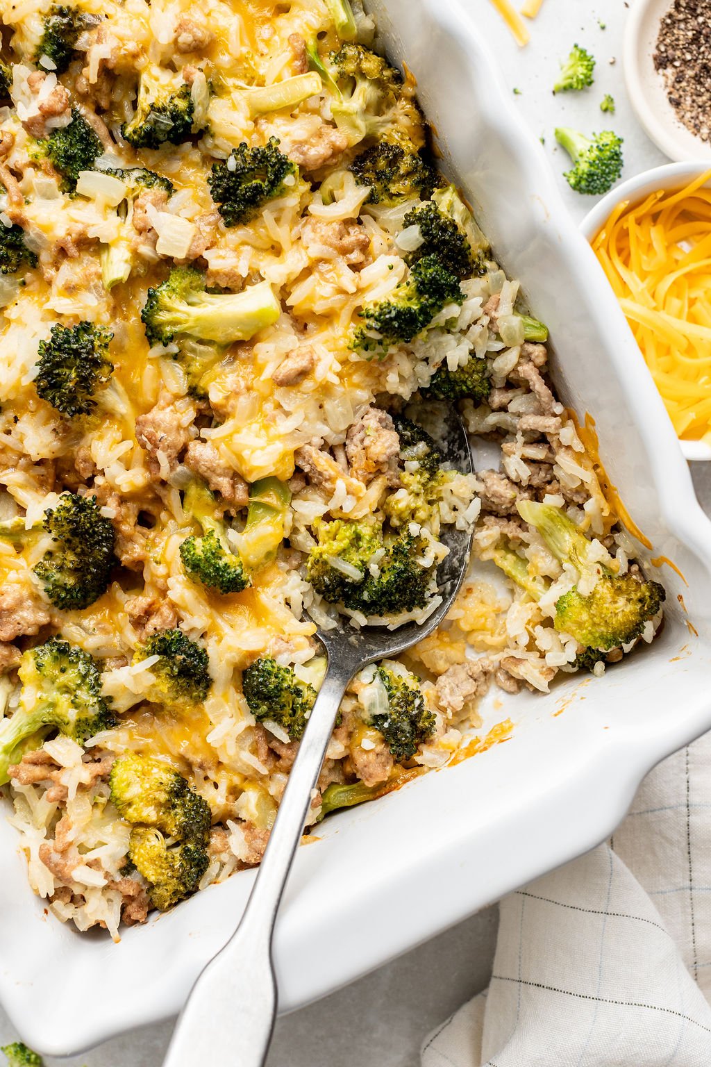 A casserole dish containing a baked mixture of rice, broccoli, ground meat, and melted cheddar cheese with a serving spoon inside.