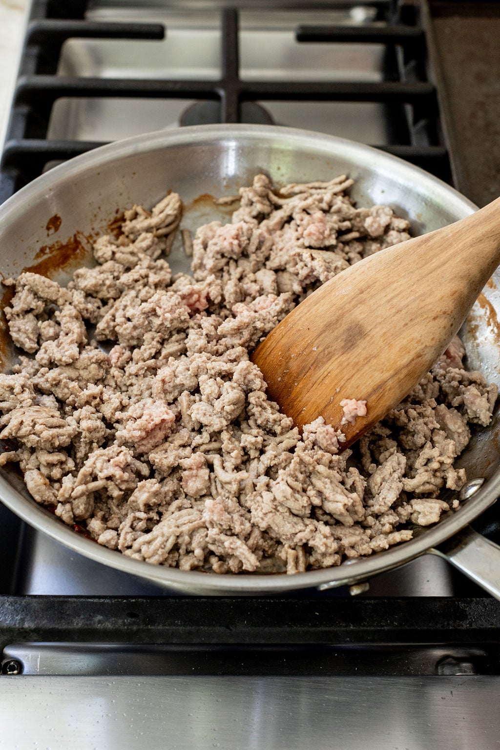 Ground meat is being browned in a skillet on a stovetop, stirred with a wooden spatula.
