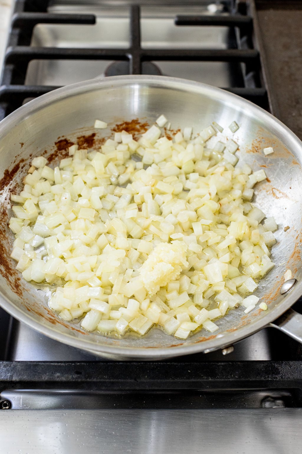 Diced onions and minced garlic are saut&eacute;ing in a stainless steel skillet on a stovetop.