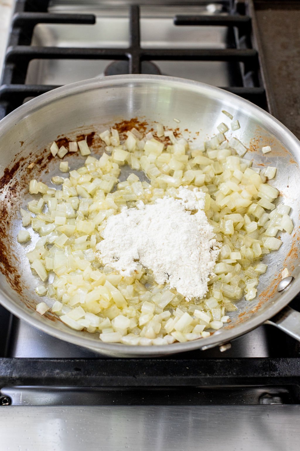 Chopped onions saut&eacute;ing in a stainless steel pan with a heap of flour added on top, placed on a gas stove.