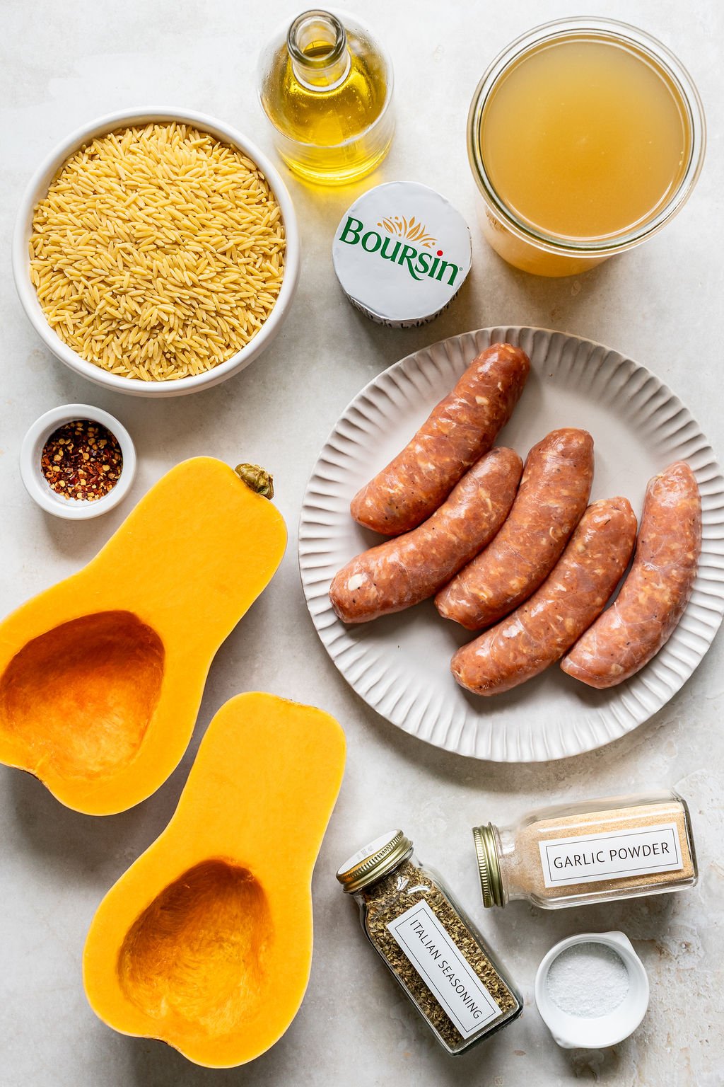 Ingredients for a recipe displayed on a table, including butternut squash halves, raw sausage, orzo, Boursin cheese, broth, oil, red pepper flakes, Italian seasoning, and garlic powder.