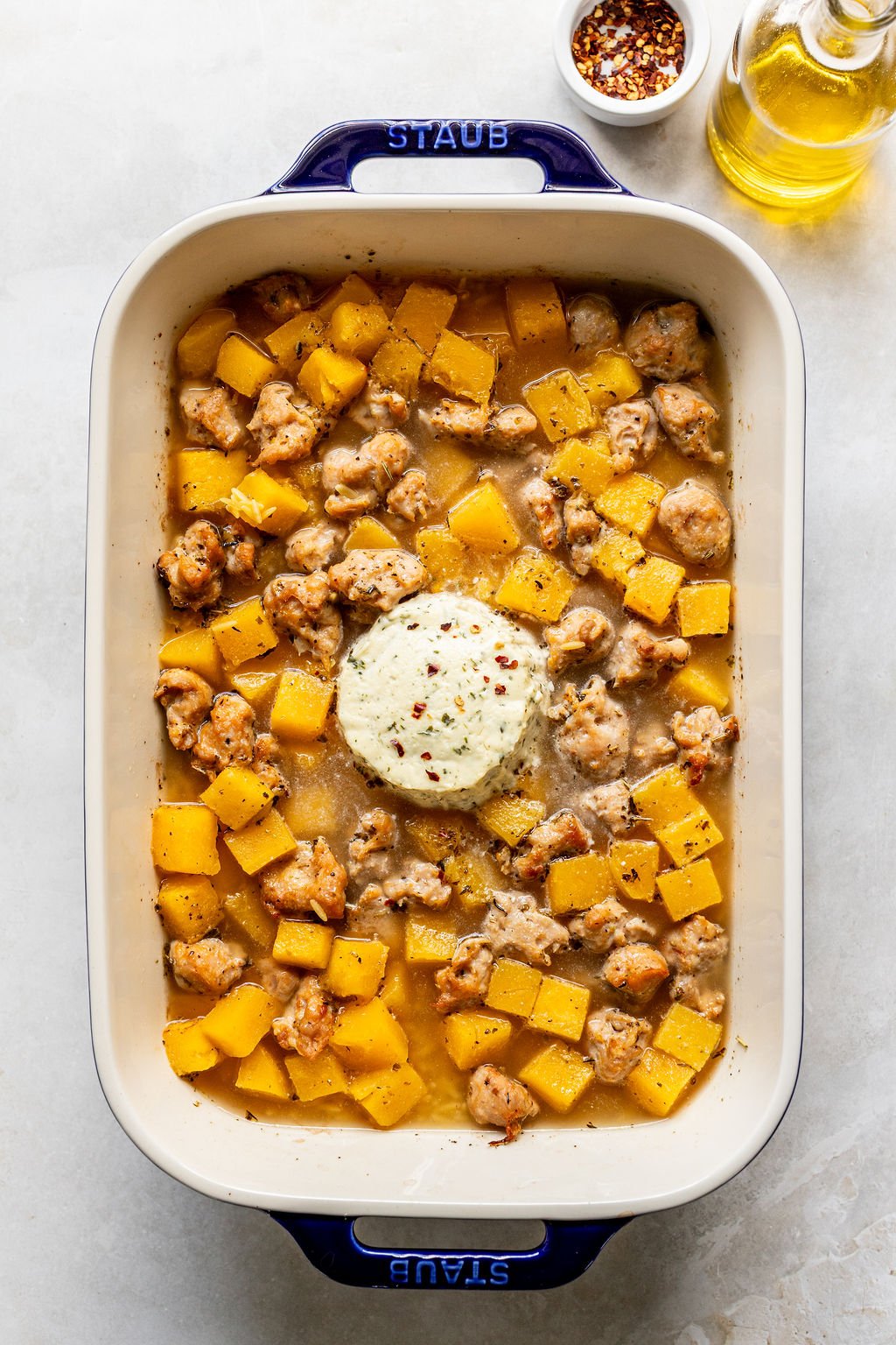 Rectangular baking dish with cooked sausage, diced butternut squash, and a round pat of herbed butter in the center. Small bowl of red pepper flakes and bottle of oil nearby.