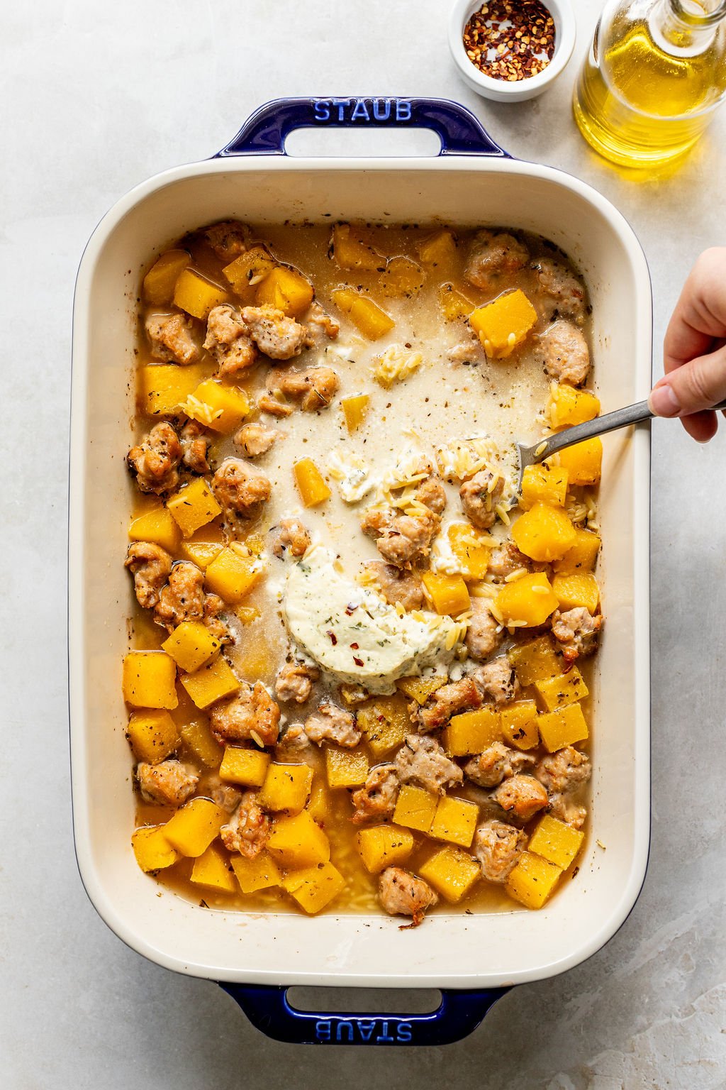 A baking dish with cooked sausage and diced butternut squash, topped with a dollop of herbed cream; a hand holds a spoon.