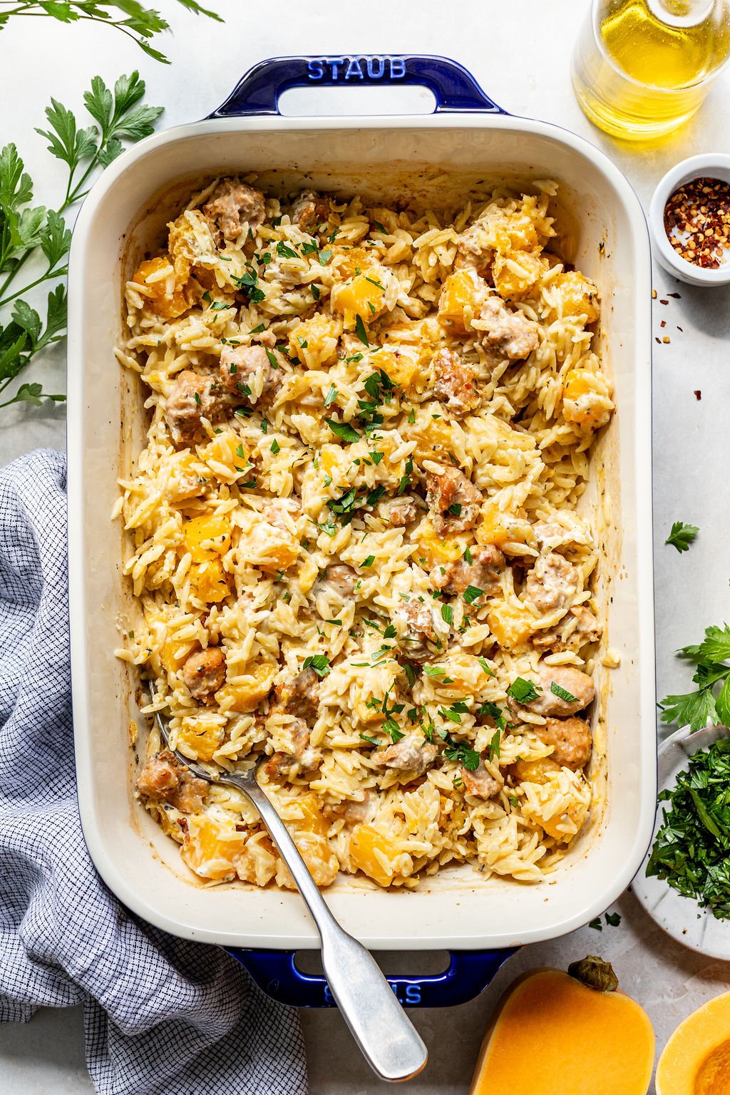 A baked casserole dish filled with orzo pasta, chunks of butternut squash, and pieces of chicken, garnished with chopped parsley.