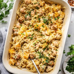 Baked casserole dish filled with orzo pasta, sausage, and chunks of butternut squash, garnished with chopped parsley. A serving spoon rests in the dish.