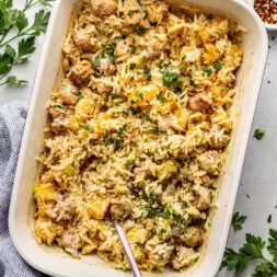 A baked casserole dish filled with orzo pasta, sausage, and butternut squash, garnished with chopped parsley, with a serving spoon inside.
