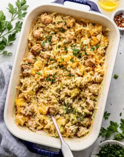 A baked casserole dish filled with orzo pasta, sausage, and butternut squash, garnished with chopped parsley, with a serving spoon inside.