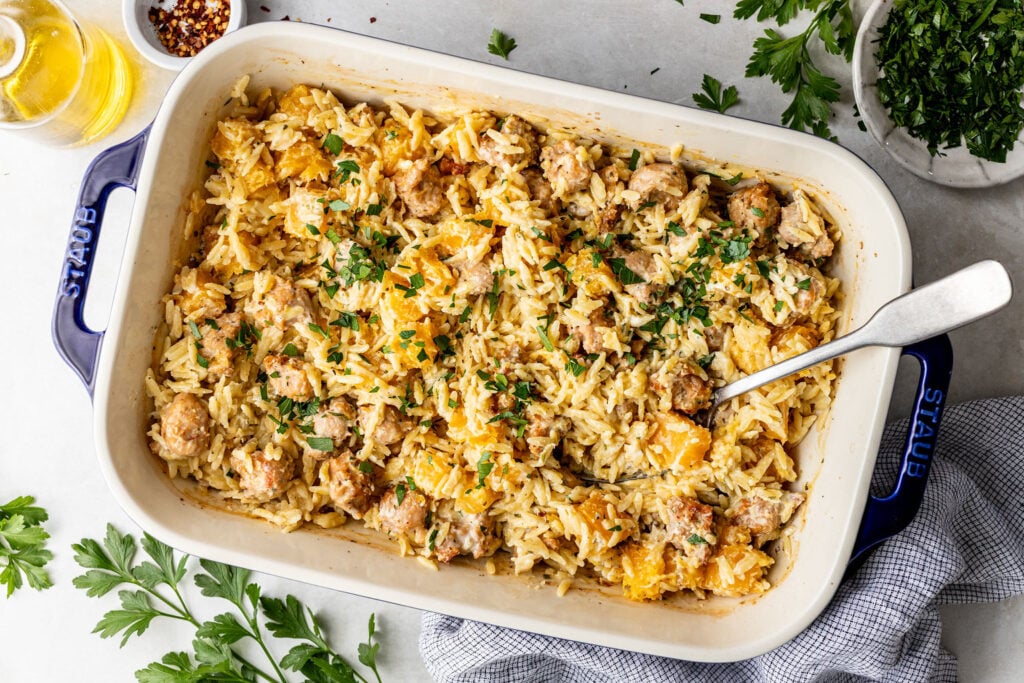 A baked casserole dish filled with orzo pasta, sausage, and butternut squash, garnished with chopped parsley, with a serving spoon inside.