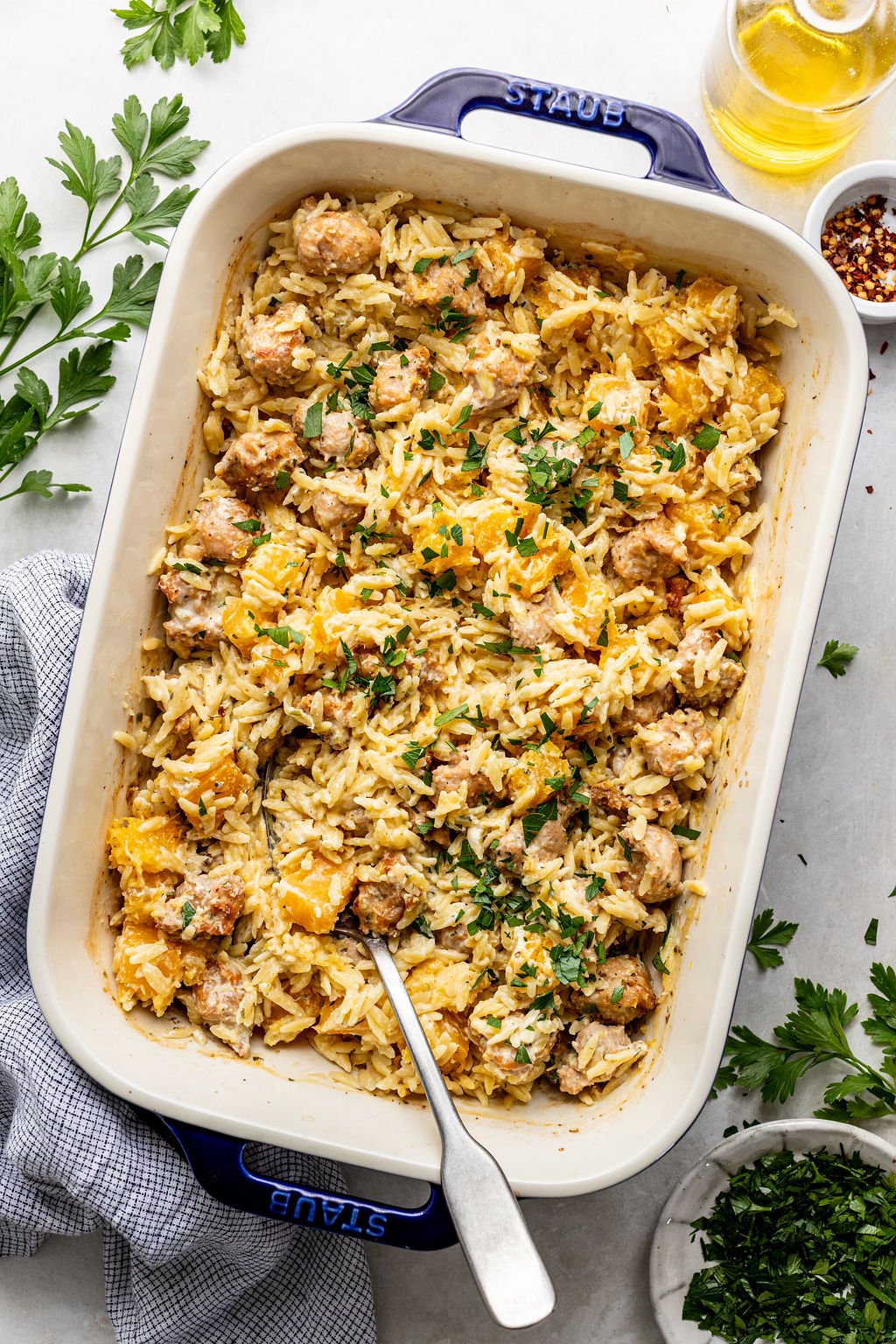 A baked casserole dish filled with orzo pasta, sausage, and butternut squash, garnished with chopped parsley, with a serving spoon inside.