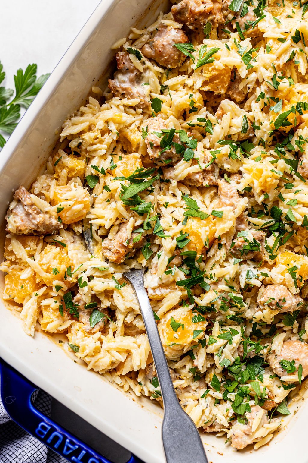 A baked orzo casserole with sausage, chunks of yellow squash, and fresh parsley, served in a white dish with a metal serving spoon.