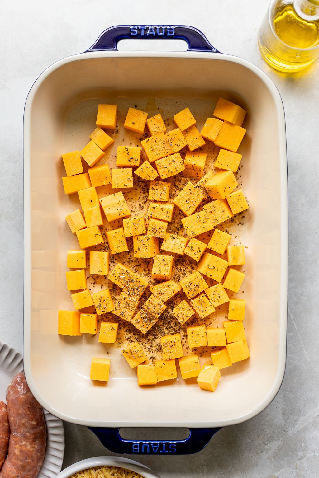 Cubed butternut squash seasoned with herbs and pepper in a rectangular baking dish, ready for roasting.