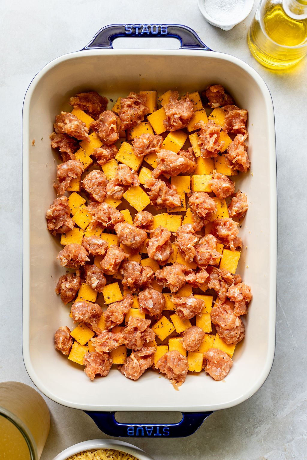 A baking dish filled with diced butternut squash and raw sausage pieces; oil and seasonings are visible on the side.
