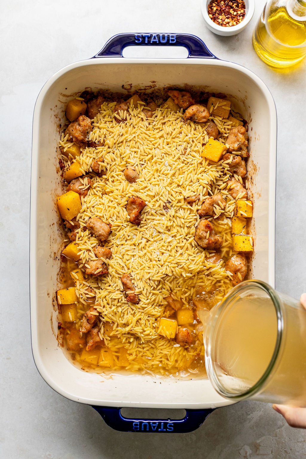 A baking dish with uncooked orzo, chicken, and squash pieces, as broth is being poured in; a small bowl of chili flakes and a bottle of oil are nearby.