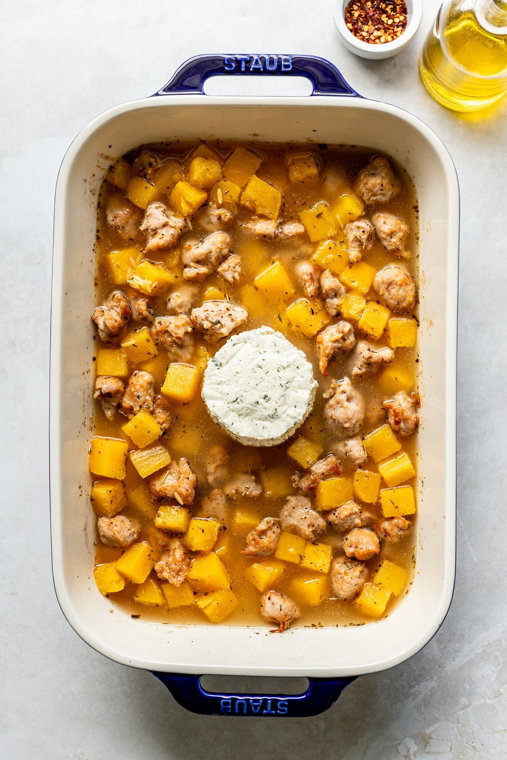 A baking dish filled with cubed squash, pieces of sausage, broth, and a whole round of herbed cheese in the center.
