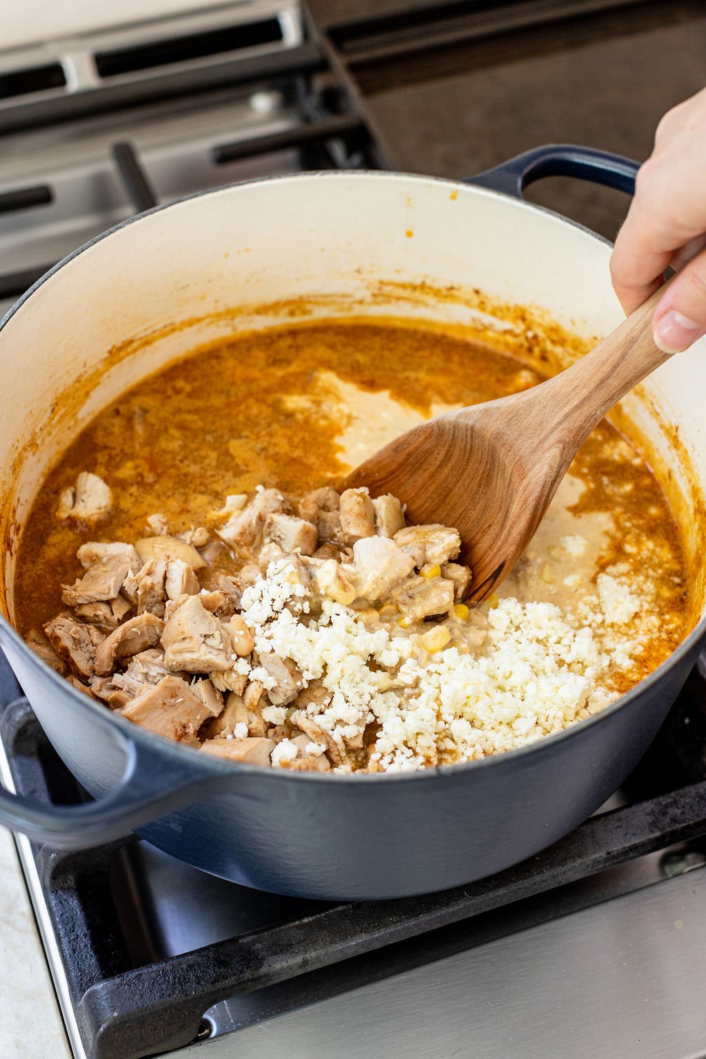 A hand stirs cooked chicken and crumbled cheese into a pot of simmering stew on a stovetop.