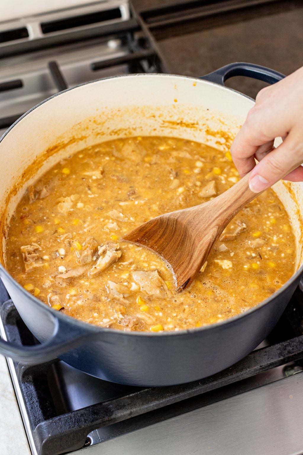 A hand stirs a pot of soup containing shredded chicken, corn, and broth on a stovetop with a wooden spoon.