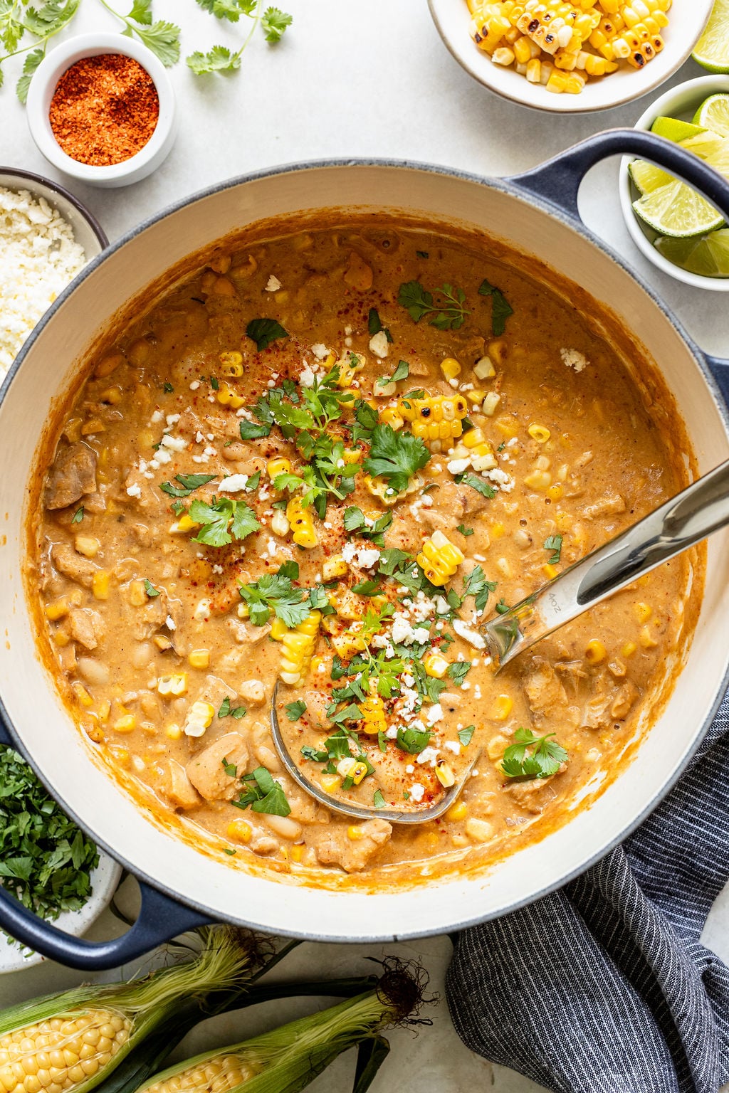 A pot of creamy corn chowder garnished with corn, cilantro, cheese, and spices, with a ladle inside; fresh corn and toppings are arranged nearby.