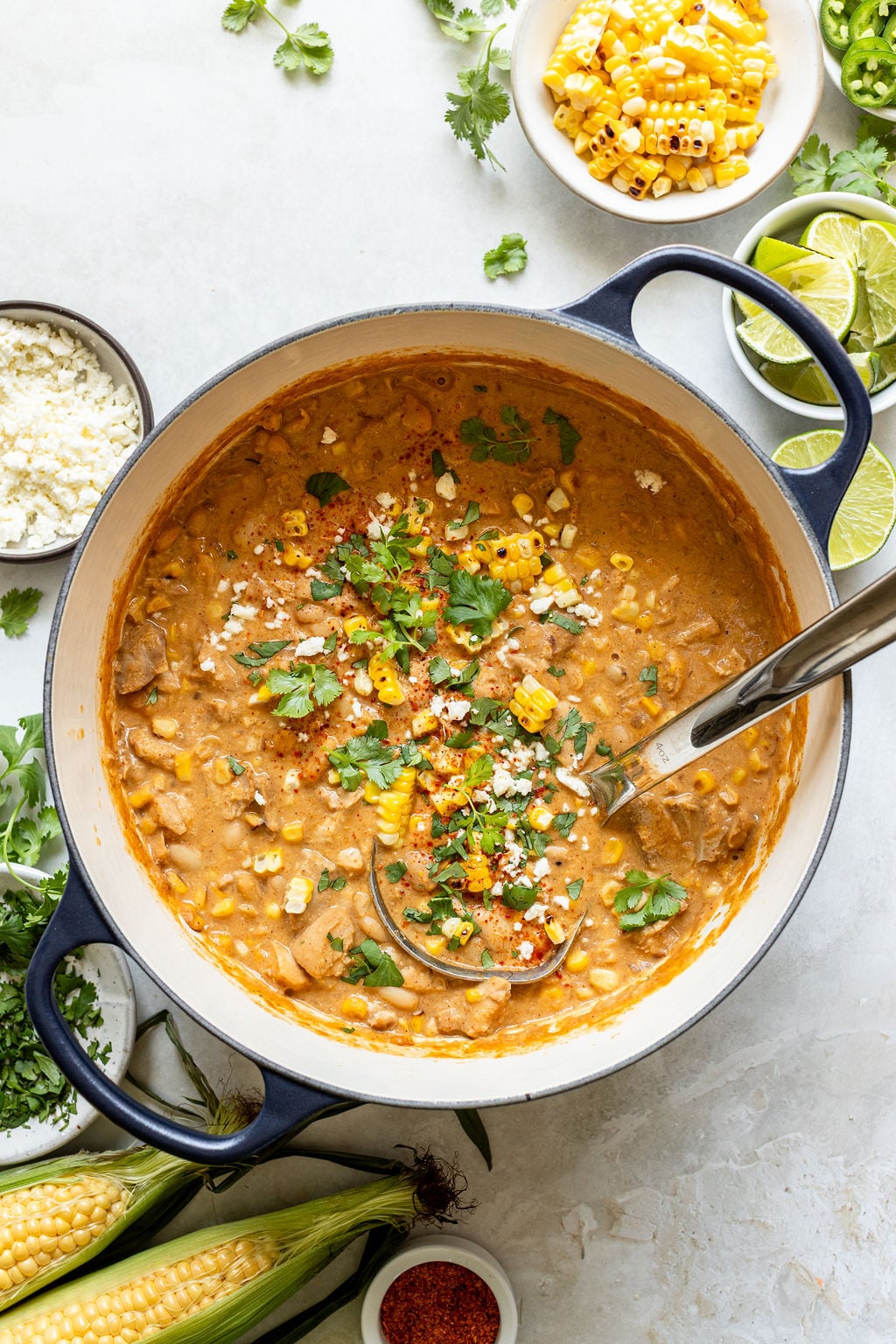 A pot of creamy soup garnished with cilantro, grilled corn, and cheese is surrounded by fresh corn, lime wedges, cilantro, cheese, and seasonings on a white surface.