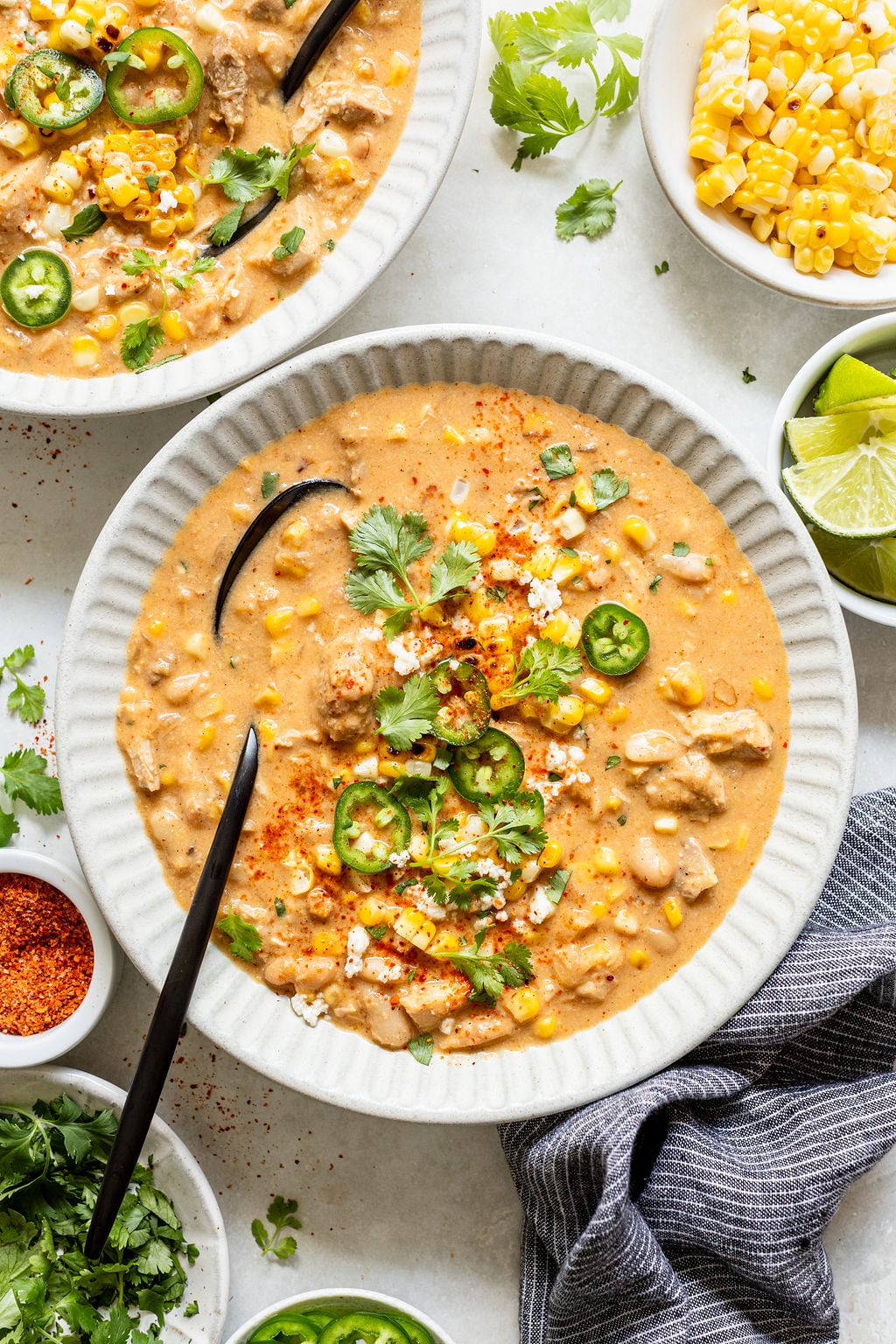 A bowl of creamy soup with corn, white beans, chicken, jalape&ntilde;o slices, cilantro, and chili powder, surrounded by bowls of toppings like lime wedges, corn, cilantro, and jalape&ntilde;os.