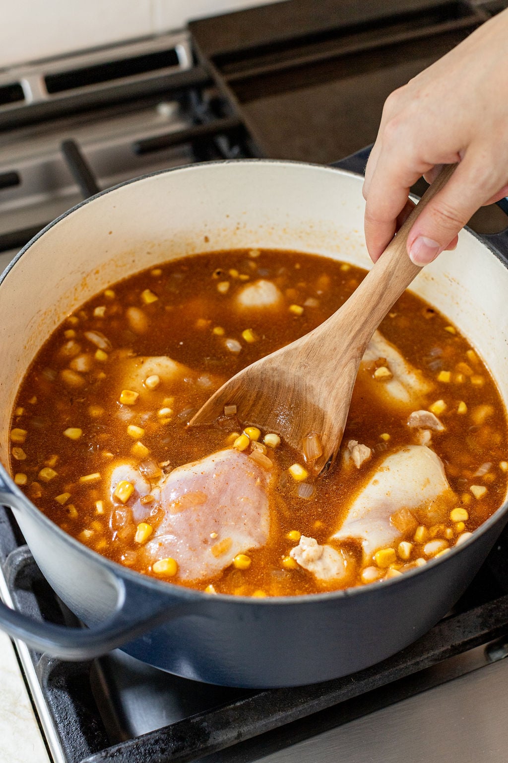 A hand stirs a pot of stew containing raw chicken, corn, beans, and broth on a stovetop with a wooden spoon.