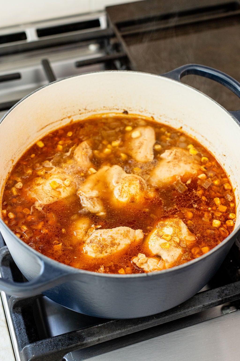A pot of chicken and corn soup simmering on a stove, with visible broth, chicken pieces, and corn kernels in a Dutch oven.