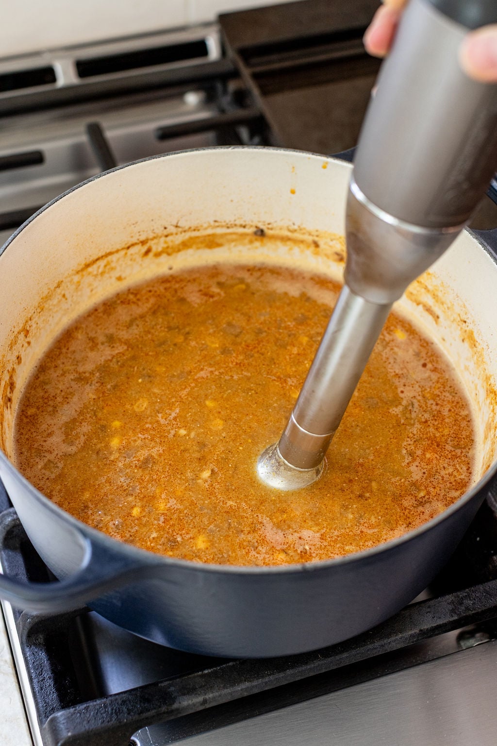 A hand uses an immersion blender to blend soup in a large blue pot on a stove.