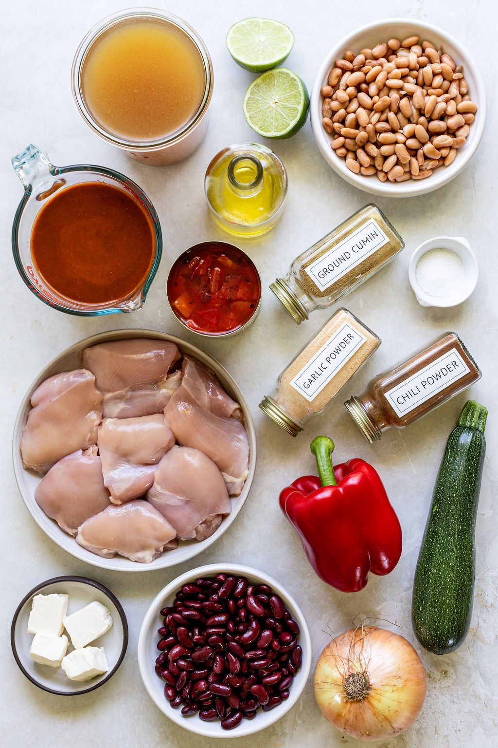 Chicken thighs, beans, broth, enchilada sauce, spices, vegetables, and oil arranged on a light surface.