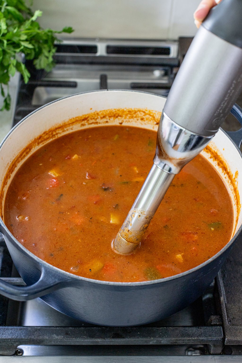 An immersion blender is blending a pot of tomato-based soup with vegetable chunks on a stovetop.
