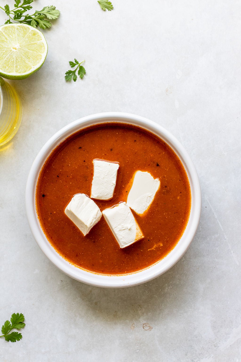 A white bowl of tomato soup topped with four cubes of cream cheese, placed on a light surface.