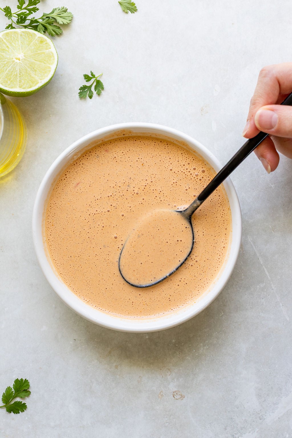 A hand holds a spoon over a bowl of creamy light-brown sauce.