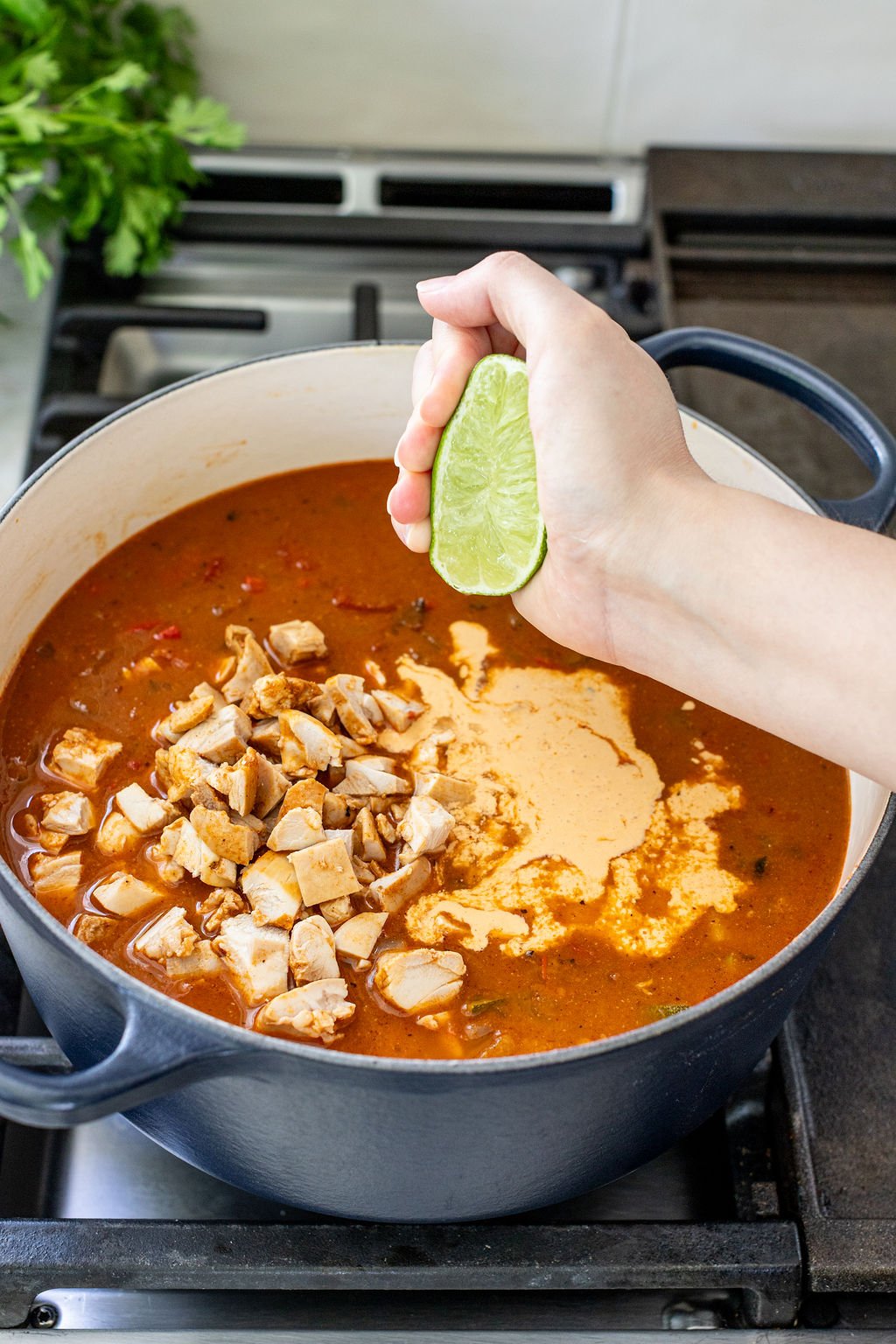 A hand squeezes a lime half over a pot of soup with chopped chicken and a creamy, reddish broth on a stovetop.
