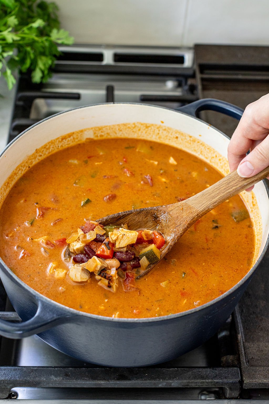 A person stirs a pot of vegetable and bean soup with a wooden spoon on a stovetop.