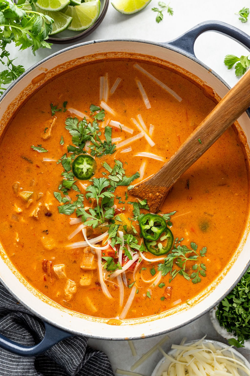 A pot of creamy orange soup garnished with shredded cheese, sliced jalape&ntilde;os, and fresh cilantro, with a wooden spoon inside. Lime wedges and herbs are nearby.