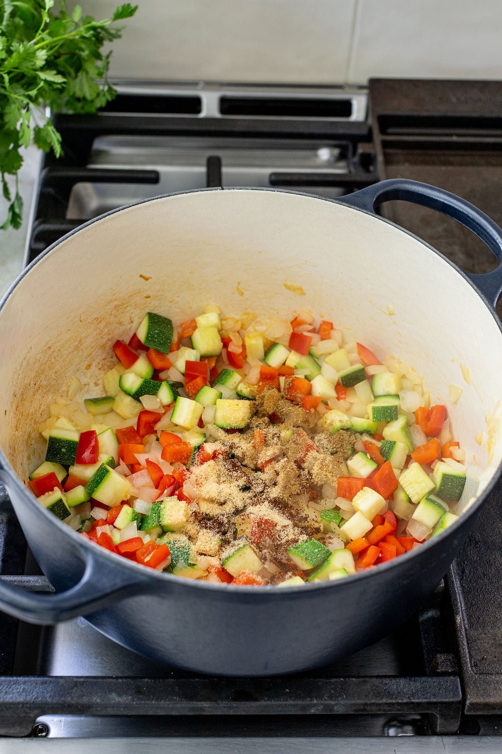 Diced zucchini, red bell pepper, and onions with spices cooking in a large pot on a stovetop.