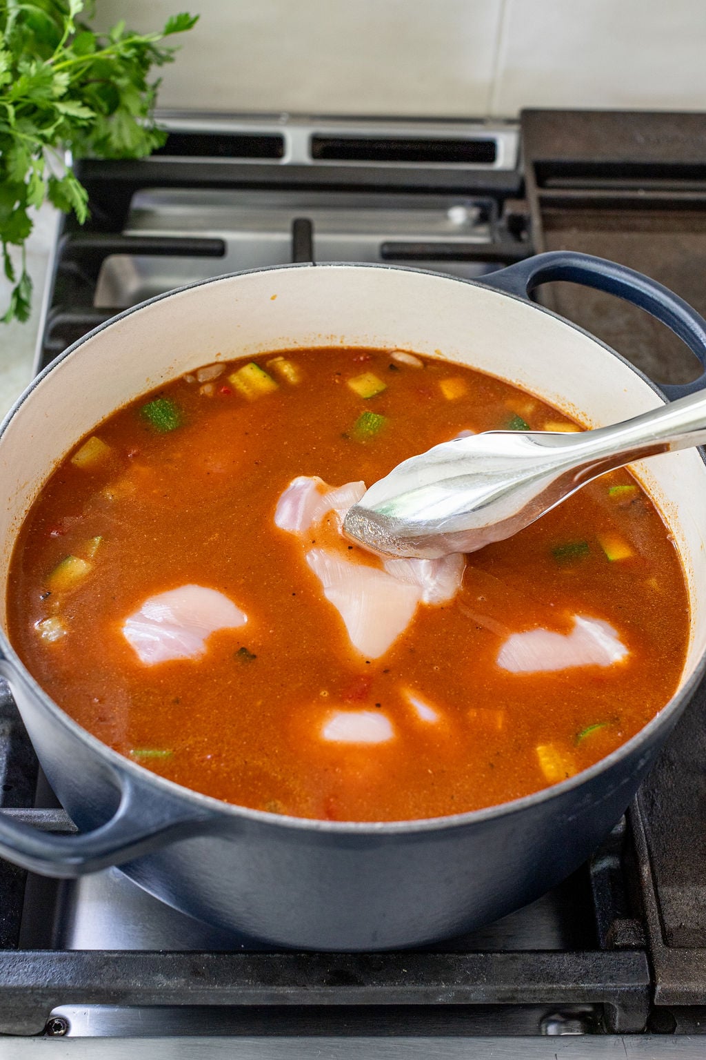 A pot of tomato-based soup with vegetables is on a stovetop, and raw chicken pieces are being added with metal tongs.
