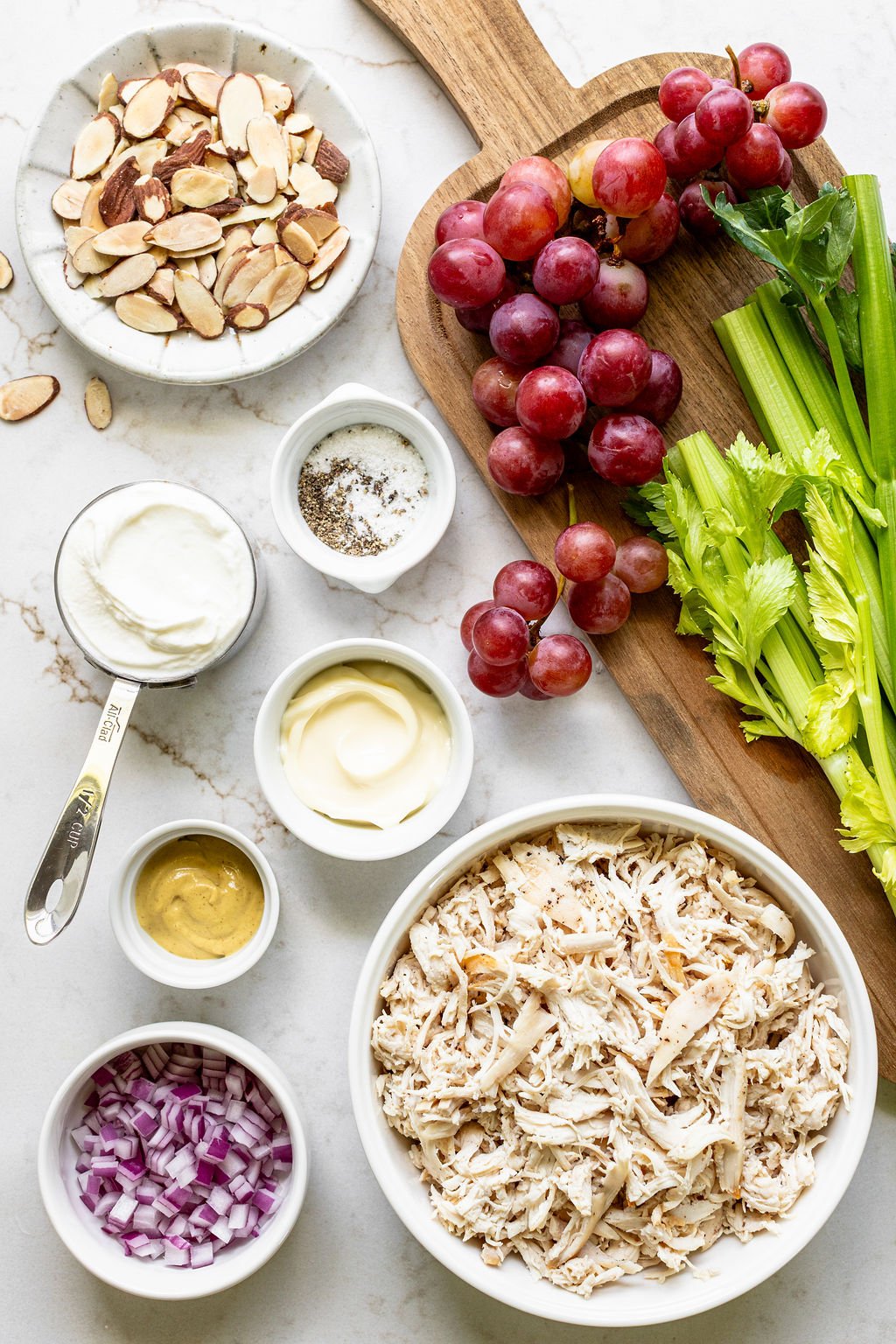 Ingredients for chicken salad arranged on a countertop, including shredded chicken, sliced almonds, red onion, mustard, yogurt, mayonnaise, grapes, celery, salt, and pepper.