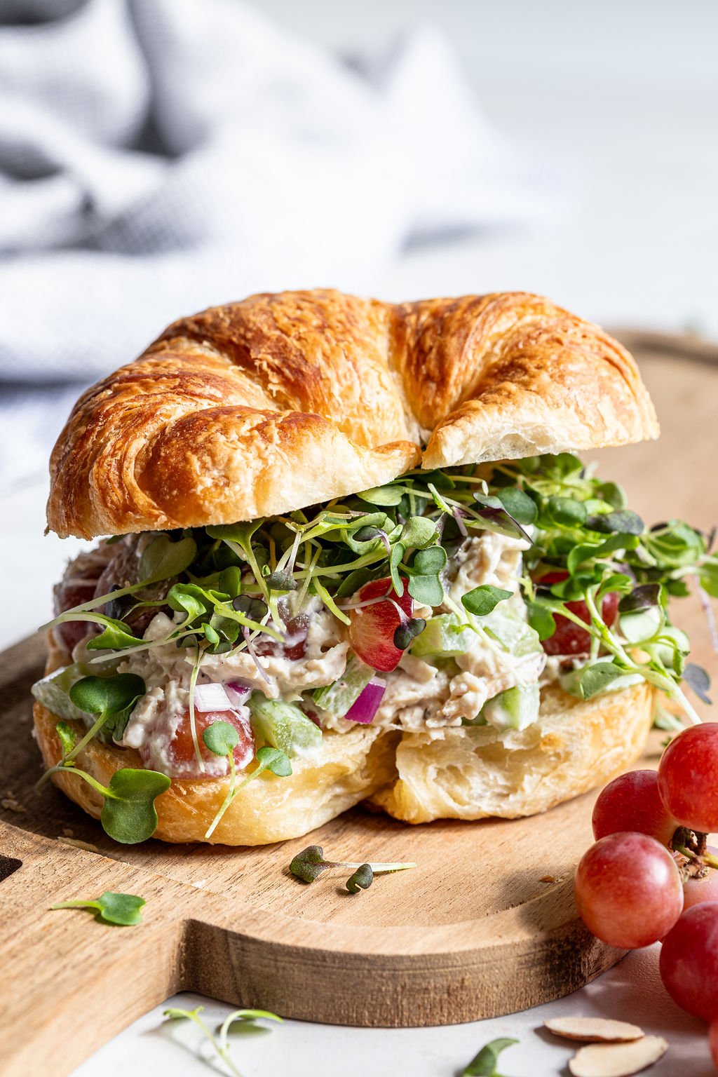 A croissant sandwich filled with chicken salad, red onions, grapes, and leafy greens sits on a wooden board with red grapes nearby.