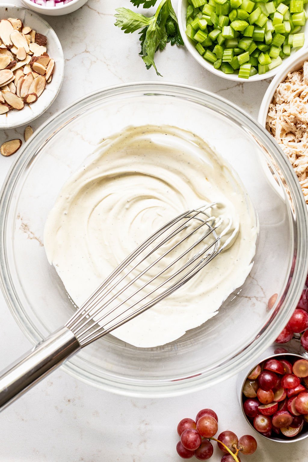 A glass bowl of creamy dressing with a whisk, surrounded by bowls of sliced almonds, chopped celery, shredded chicken, red grapes, and parsley on a marble surface.