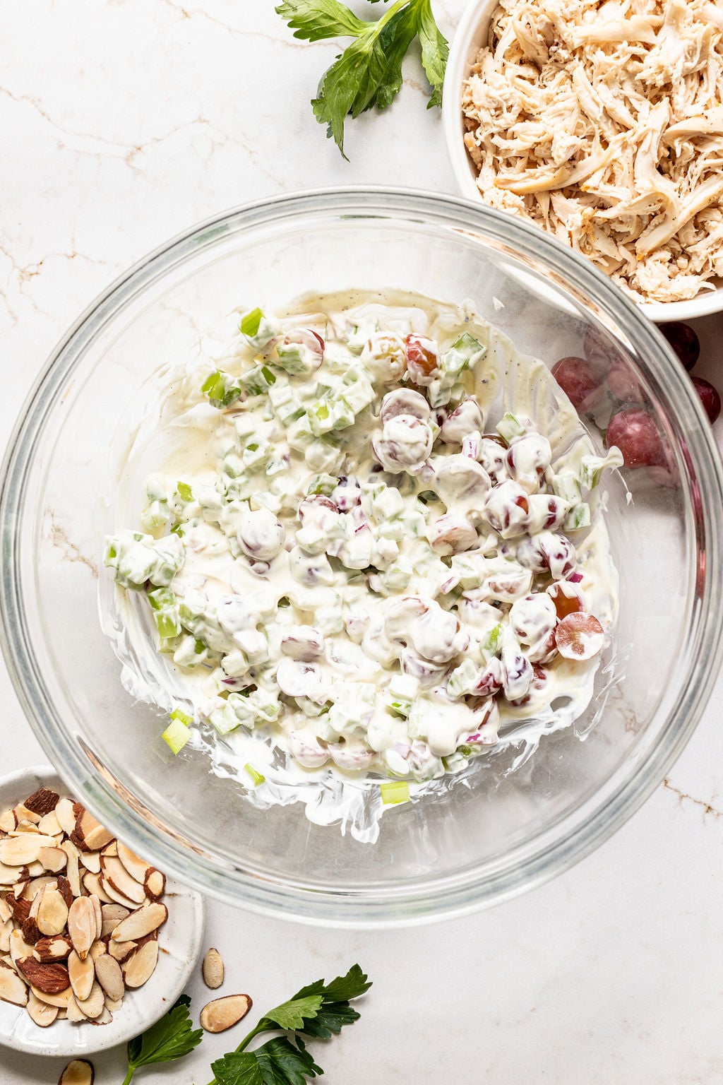 A glass bowl with creamy chicken salad mixture, surrounded by a bowl of shredded chicken, a plate of sliced almonds, and fresh parsley on a marble countertop.