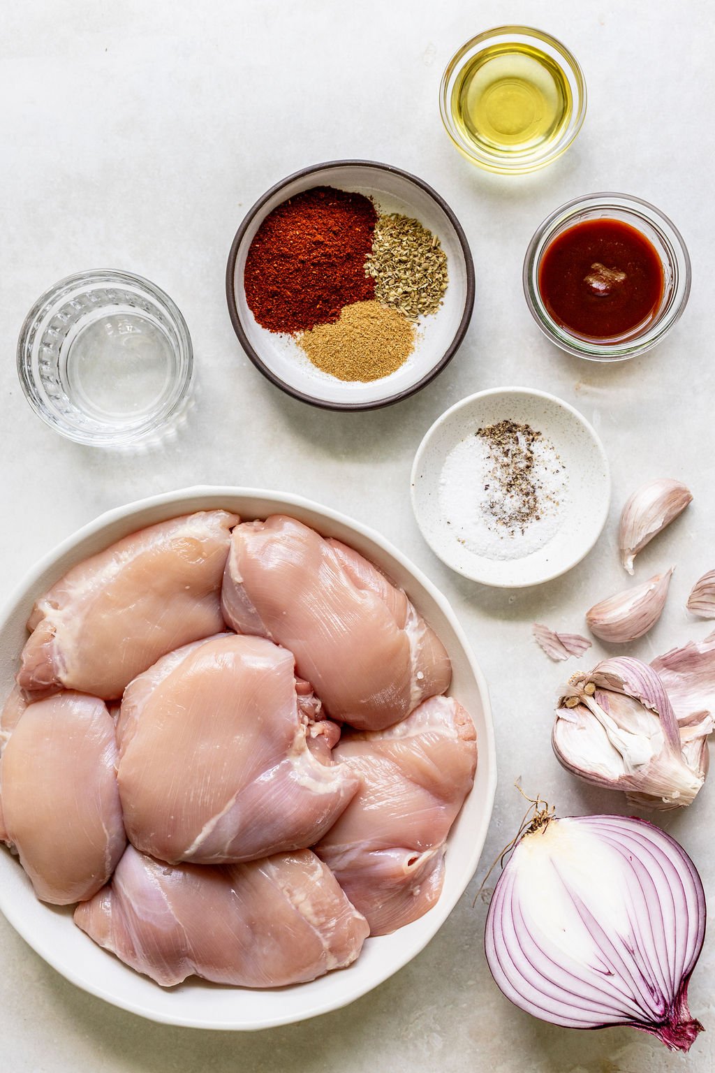 Raw chicken thighs in a bowl, surrounded by seasonings, olive oil, hot sauce, water, salt, garlic cloves, and a halved red onion on a white surface.