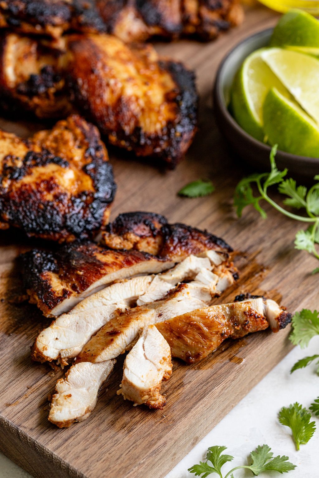 Sliced grilled chicken breast on a wooden board, with charred edges, garnished with cilantro and lime wedges in a small bowl.