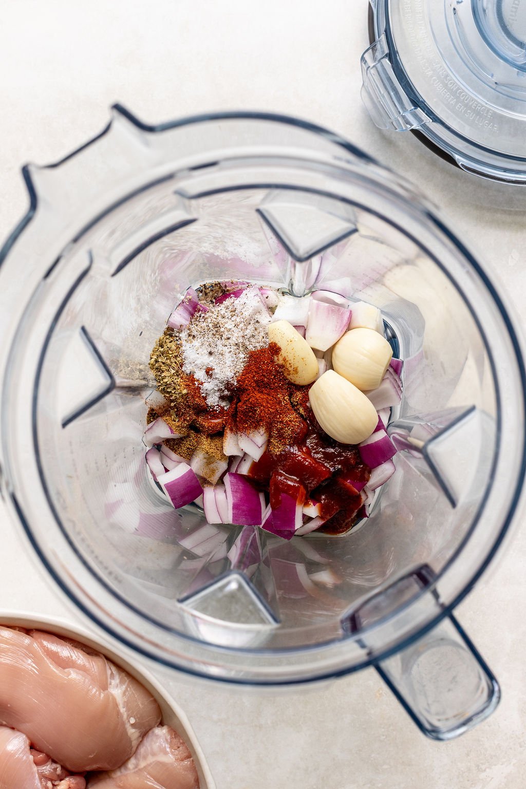 Chopped red onion, garlic cloves, spices, and seasonings are placed in a blender; raw chicken is visible in a bowl nearby.