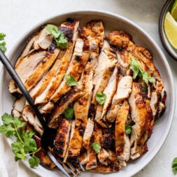 A plate of sliced grilled chicken garnished with cilantro, with a fork on the side. Rice, lime wedges, and more cilantro are nearby.