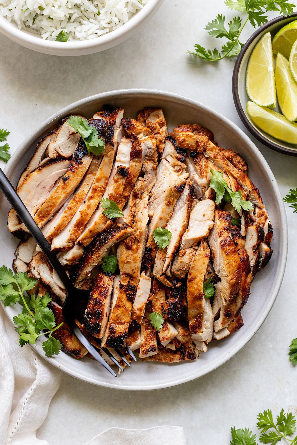 A plate of sliced, grilled chicken garnished with cilantro, served with lime wedges and a bowl of rice on the side.