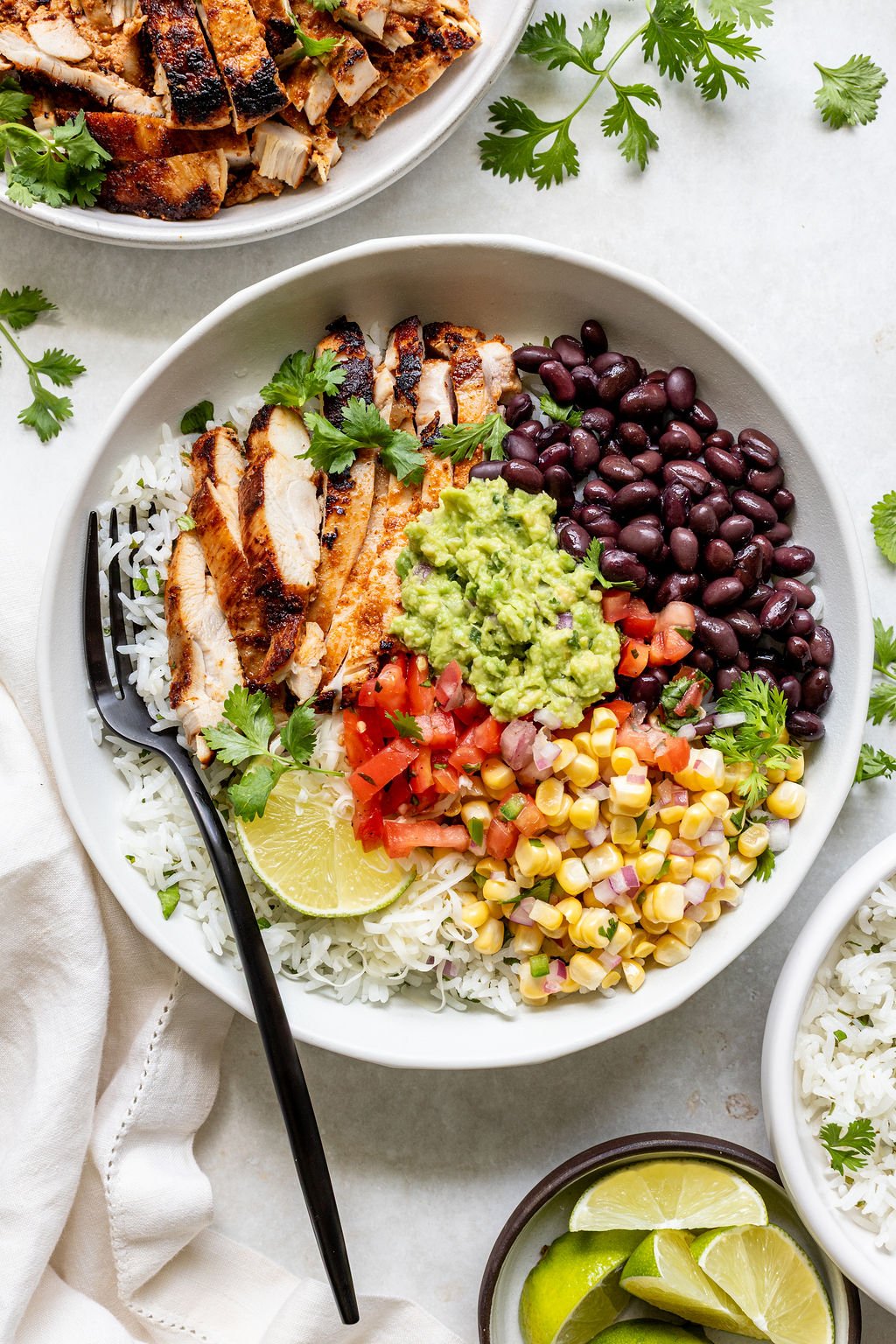 A bowl with sliced grilled chicken, black beans, guacamole, diced tomatoes, corn, cilantro, rice, and a lime wedge, with a black fork on the side.